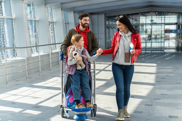 Ganzkörperansicht der brünetten Frau, die ihr hübsches Kind ansieht und ihre Hand hält, während sie am Flughafen spazieren geht. Vater lächelt vergnügt. Glückliche Familie mit Koffern im Flughafenkonzept