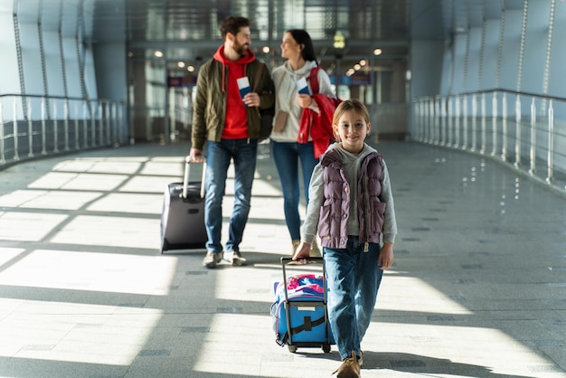 Ganzkörperansicht der glücklichen jungen Familie von Mutter, Vater und Tochter, die mit Koffern durch die Halle geht und auf den Flug am Flughafen wartet. Reisekonzept