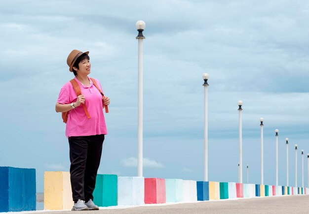 Ganzkörperansicht einer lächelnden asiatischen Touristin, die seitlich auf der Brücke am Aussichtspunkt auf das Meer blickt