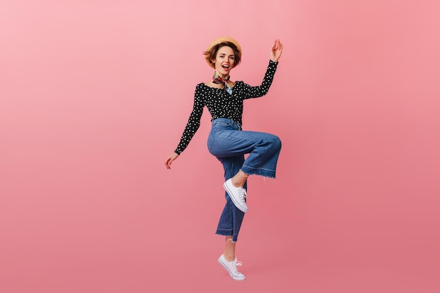 Ganzkörperansicht einer springenden Frau mit Strohhut Studioaufnahme eines Mädchens in Vintage-Jeans, das auf rosafarbenem Hintergrund tanzt