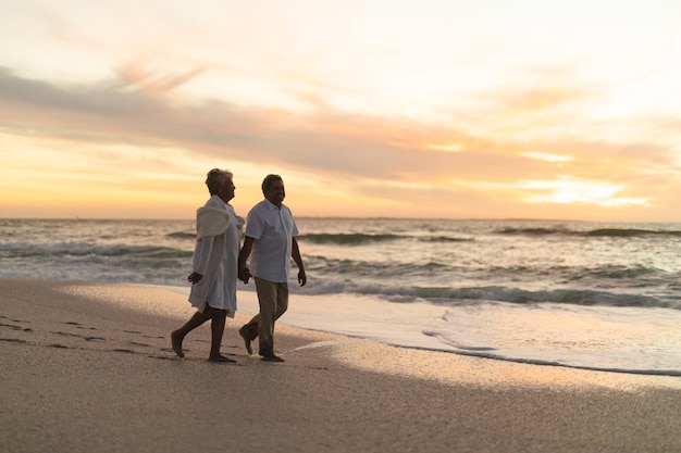 Ganzkörperansicht eines älteren, gemischtrassigen Paares, das gemeinsam den Sonnenuntergang verbringt, während es am Strand gegen den Himmel spaziert