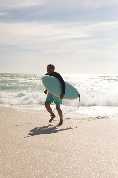 Ganzkörperansicht eines biracial älteren Mannes mit Surfbrett, der am Strand gegen den Himmel läuft