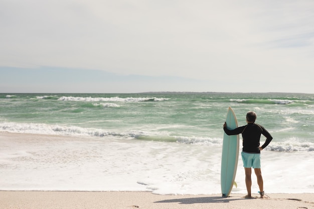 Ganzkörperansicht eines biracial älteren Mannes mit Surfbrett, der an einem sonnigen Tag am Strand steht