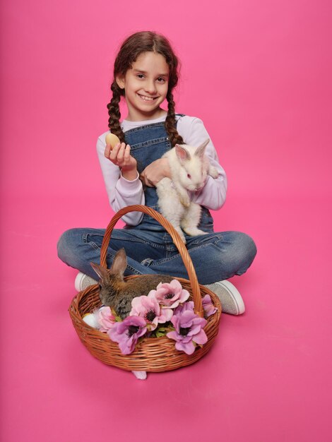 Ganzkörperansicht eines entzückenden kleinen Mädchens, das auf dem Boden sitzt und ein kleines Kaninchen in den Händen hält, das isoliert auf einem rosa Hintergrund in die Kamera blickt Vertikale Ansicht