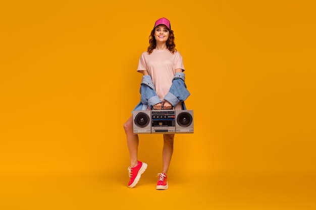 Ganzkörperansicht von ihr, sie ist ein hübsches, hübsches, modisches, fröhliches, fröhliches Mädchen, das einen coolen Look trägt, der Boombox einzeln auf hell leuchtendem, leuchtend gelbem Hintergrund trägt