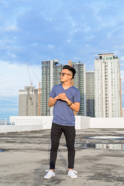 Ganzkörperaufnahme des jungen gutaussehenden multiethnischen Mannes mit Sonnenbrille gegen Blick auf die Stadt