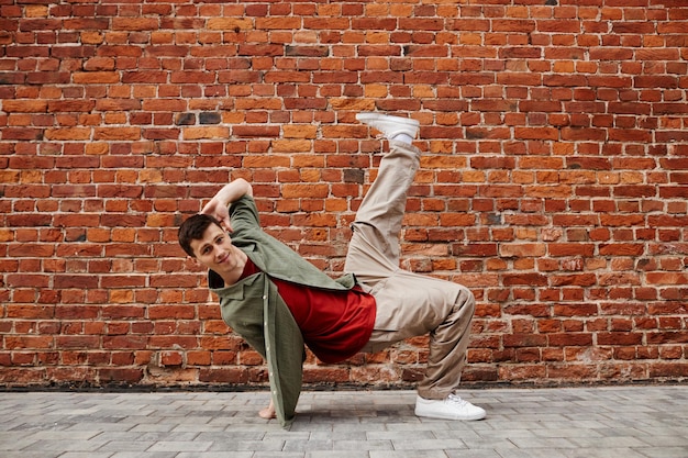 Ganzkörperaufnahme eines jungen Mannes, der Hiphop-Tanz-Pose macht und in die Kamera lächelt, gegen eine Backsteinmauer-Kopie