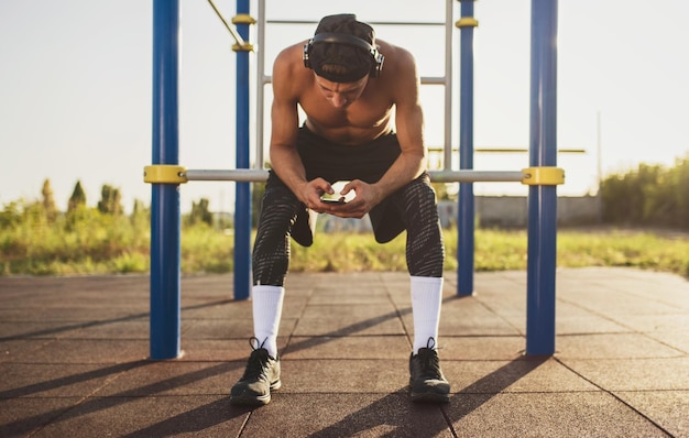 Ganzkörperbild eines athletischen, hemdlosen, muskulösen jungen Mannes, der sich nach dem Training auf dem Trainingsgelände im Freien entspannt Schöner Sportler, der sein Smartphone für Sportanwendungen mit Kopfhörern verwendet