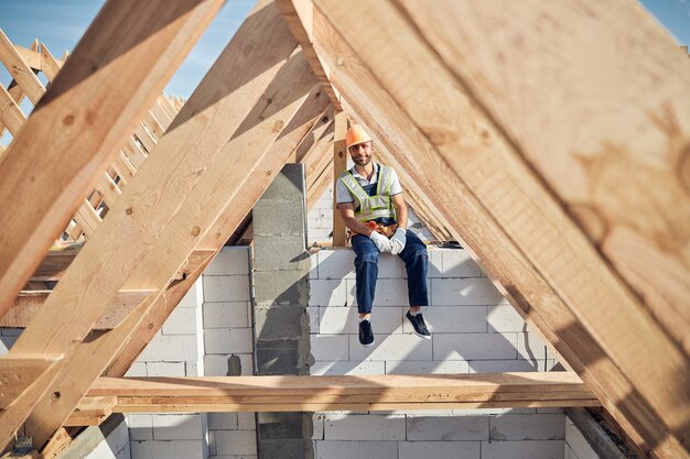 Ganzkörperfoto eines Architekten, der einen Schutzhelm trägt, während er auf einer Gebäudewand sitzt und in die Kamera schaut