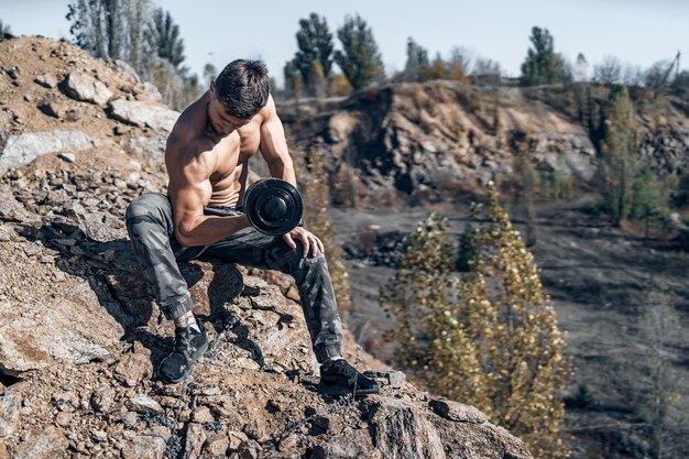 Ganzkörperfoto eines Bodybuilders mit Metallhantel in der Hand Shirtless Bodybuilder Rocks on the background