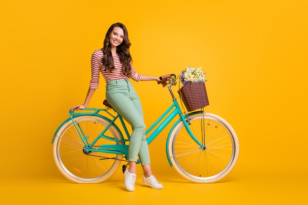 Ganzkörperfoto eines hübschen Mädchens, das auf einem Fahrrad sitzt und einen Blumenkorb lächelt, isoliert auf leuchtend gelbem Hintergrund