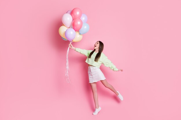 Ganzkörperfoto eines überraschten, schockierten, verrückten, fröhlichen Mädchens, das viele Luftballons hält, die Luft fliegen beeindruckt, genießen, sich freuen, weiße Pullover-Turnschuhe tragen, isoliert pastellfarbener Hintergrund