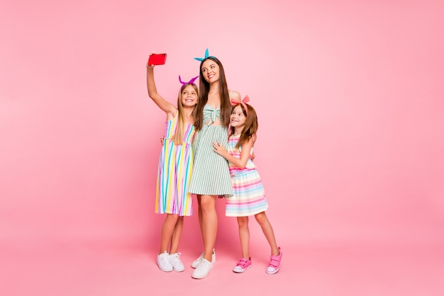 Ganzkörperfoto von niedlichen drei Personen mit langen brünetten blonden Haaren, die Selfie tragen Rockkleid Stirnbänder lokalisiert über rosa Hintergrund machen