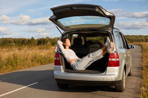 Ganzkörperporträt einer entspannten Frau, die im offenen Kofferraum eines Autos sitzt und sich in der Sonne aalt. Frau mit weißem Hemd und Jeans. Schöne Frau, die allein reist