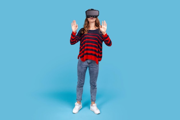 Ganzkörperporträt einer Frau, die einen gestreiften Freizeitpullover trägt, neue Technologien entdeckt, in einem Virtual-Reality-Headset steht, futuristische 3D-Vision. Studioaufnahme im Innenbereich isoliert auf blauem Hintergrund.
