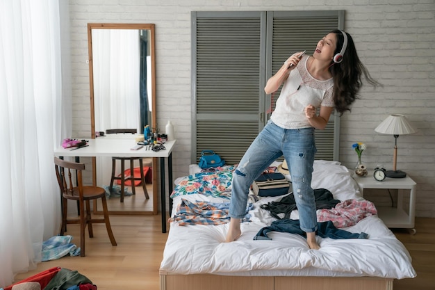 Ganzkörperporträt einer jungen, lässigen Frau mit Kopfhörern, die Musik hört und drinnen auf dem Bett tanzt, während sie im Schlafzimmer Kleidung trägt, während sie Koffer packt. fröhliche frau, die handy hält