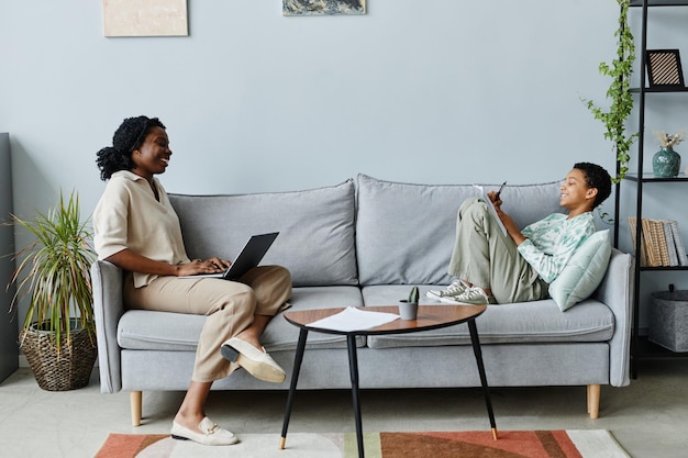 Ganzkörperporträt einer lächelnden Tutorin, die einem schwarzen Teenager hilft, zu Hause auf der Couch zu lernen...