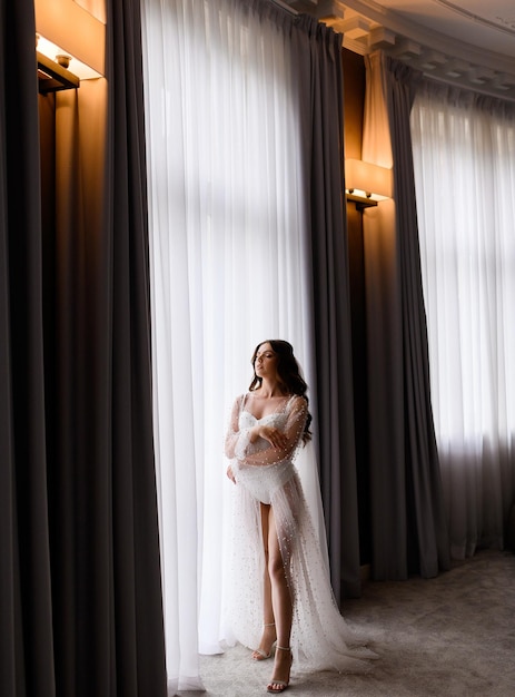 Ganzkörperporträt einer sinnlichen Frau in leichtem Hochzeitskleid in der Nähe eines großen Fensters mit geschlossenen Augen Brautfrau genießt einen besonderen Moment, der in einer Luxuswohnung posiert