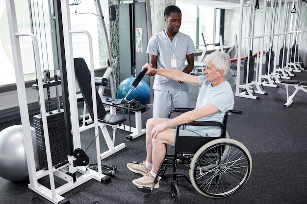 Ganzkörperporträt eines älteren Mannes, der im Fitnessstudio einen Rollstuhl benutzt, wobei der Rehabilitationstherapeut c unterstützt