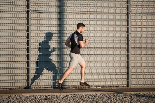 Ganzkörperporträt eines athletischen Mannes, der draußen entlang einer schönen grauen Wand läuft, muskulöser junger Läufer, der beim Joggen im Park trainiert. Sonnenuntergang