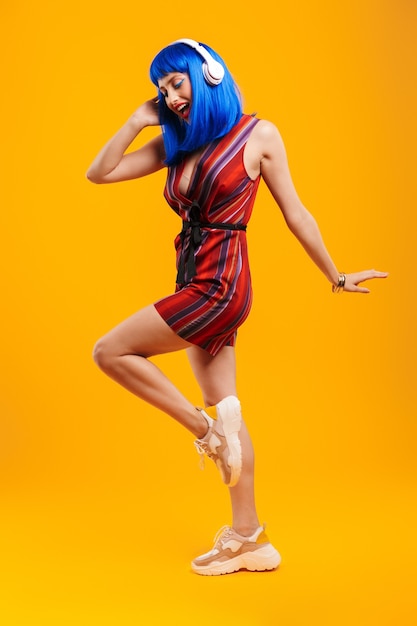 Ganzkörperporträt eines attraktiven, funky jungen Mädchens mit blauen Haaren, das ein lässiges Outfit trägt, das isoliert über eine gelbe Wand springt und Musik mit Kopfhörern hört
