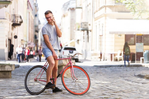 Ganzkörperporträt eines gutaussehenden jungen kaukasischen Mannes in lässiger Kleidung, der mit dem Telefon wegschaut und lächelt, während er sich auf sein Fahrrad lehnt und im Freien steht