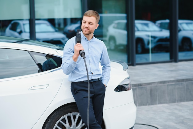 Ganzkörperporträt eines jungen gutaussehenden bärtigen Mannes in Freizeitkleidung, der an der Ladestation steht und einen Stecker des Ladegeräts für ein Elektroauto hält Öko-Elektroauto-Konzept