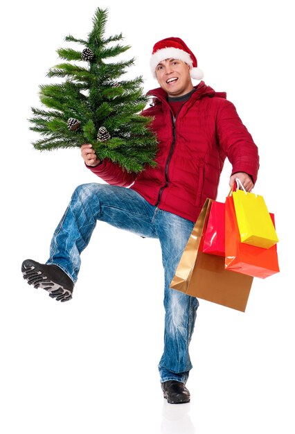 Foto ganzkörperporträt eines jungen mannes in winterkleidung und weihnachtsmütze mit einkaufstüten und künstlicher tanne isoliert auf weißem hintergrund