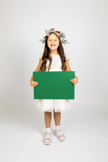 Ganzkörperporträt eines lachenden fröhlichen Mädchens in Weihnachtskranz und weißem festlichem Kleid mit Gr...