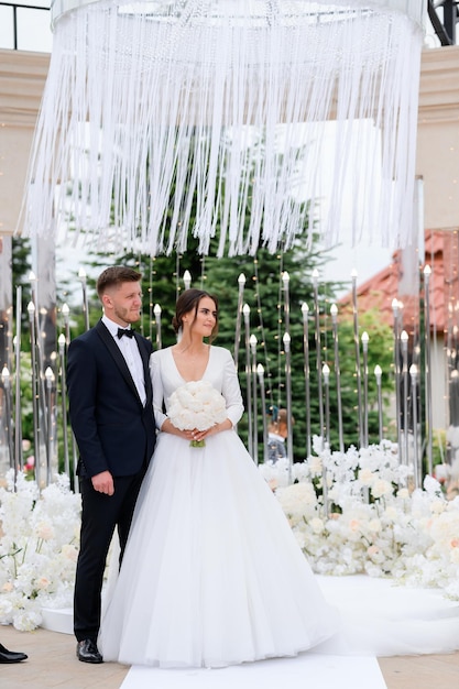 Ganzkörperporträt eines liebevollen Ehepaares, das auf einem stilvollen Hochzeitsaltar steht, mit Blumen, die Grüße hören Gut aussehender Bräutigam im schwarzen Anzug, der die Braut in einem eleganten Satinkleid umarmt