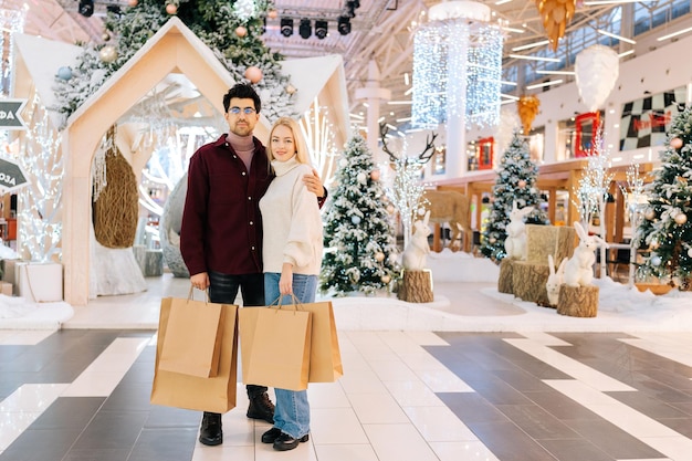 Ganzkörperporträt eines schönen jungen Paares, das Einkaufspapiertüten mit Geschenken hält und in die Kamera blickt, die in der Halle des Feier-Einkaufszentrums steht