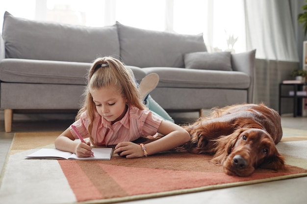 Ganzkörperporträt eines süßen blonden Mädchens, das Hausaufgaben macht, während es mit Hund auf dem Boden liegt