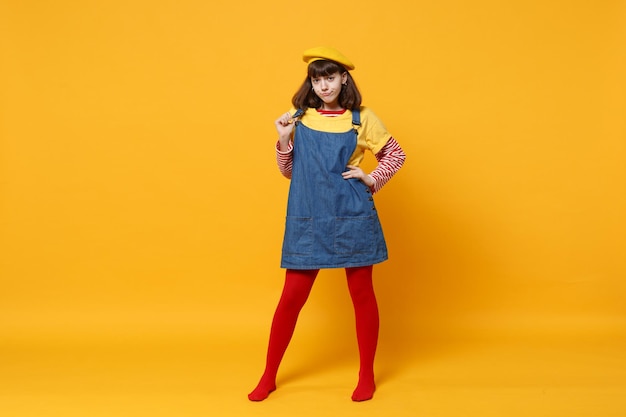 Ganzkörperporträt eines verwirrten Teenagers in französischer Baskenmütze und Denim-Sommerkleid, der mit in die Seite gestemmtem Arm steht, isoliert auf gelbem Wandhintergrund. Menschen Emotionen, Lifestyle-Konzept. Mock-up-Kopienbereich.
