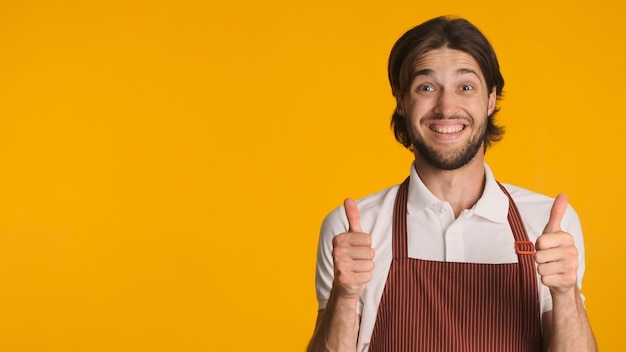 Garçom alegre vestindo avental mantendo os polegares para cima mostrando gesto aprovado na câmera sobre fundo colorido Linguagem corporal