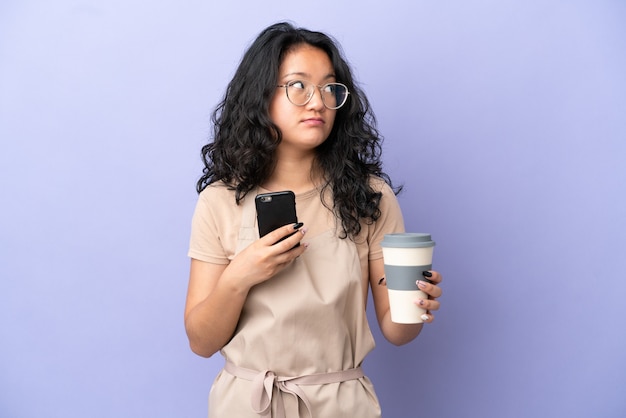 Garçom asiático de restaurante isolado em um fundo roxo segurando um café para levar e um celular enquanto pensava em algo