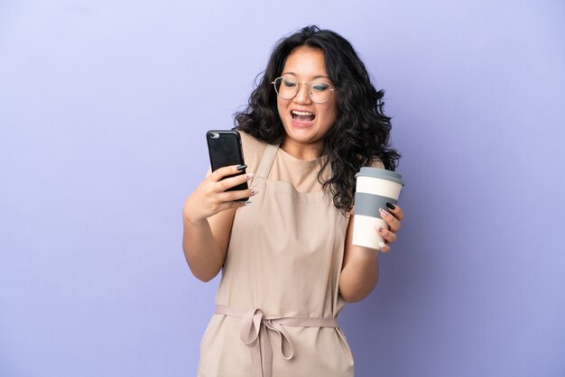 Garçom asiático de restaurante isolado em um fundo roxo segurando um café para levar e um celular