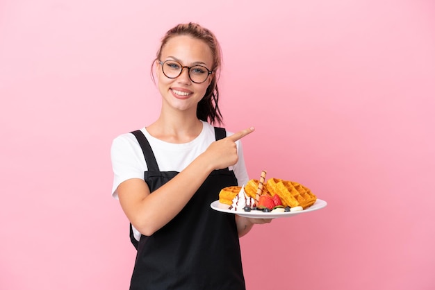 Garçom de restaurante garota russa segurando waffles isolados em fundo rosa apontando para trás