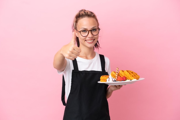 Garçom de restaurante garota russa segurando waffles isolados em fundo rosa com polegares para cima porque algo bom aconteceu