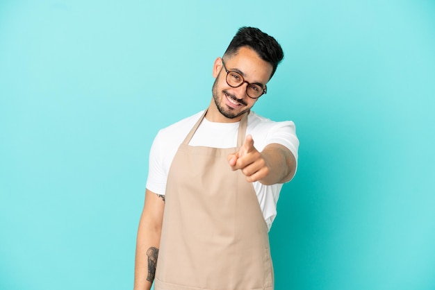 Garçom de restaurante, homem caucasiano, isolado em um fundo azul, apontando o dedo para você com uma expressão confiante