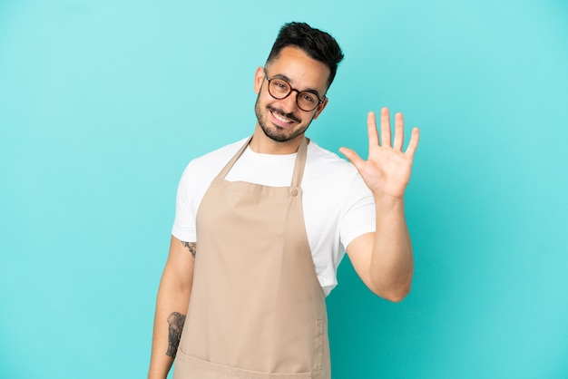 Garçom de restaurante, homem caucasiano, isolado em um fundo azul, saudando com a mão e com uma expressão feliz