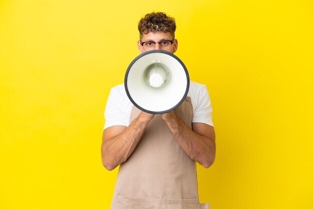 Garçom de restaurante loiro isolado em fundo amarelo gritando através de um megafone