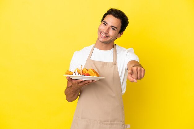 Garçom do restaurante segurando waffles sobre um fundo amarelo isolado apontando para a frente com uma expressão feliz