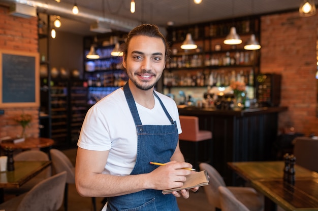 Garçom jovem e feliz com camiseta branca e avental azul escuro olhando para você com um sorriso largo e escrevendo seu pedido no restaurante