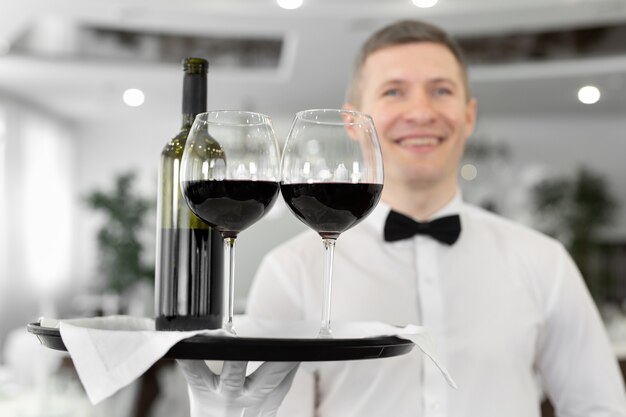 Garçom sorridente com copos de vinho tinto e uma garrafa em uma bandeja em um restaurante.