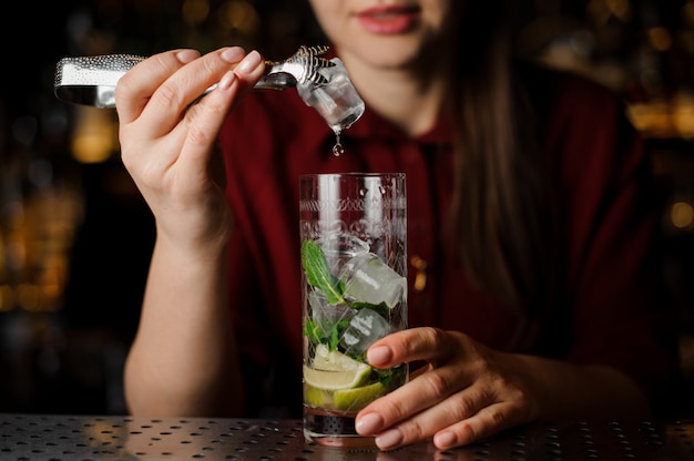 Garçonete bonita em um vestido vermelho prepara um mojito usando pinças de gelo