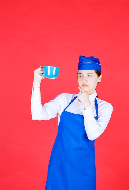 Garçonete de mulher de uniforme em pé e segurando uma xícara azul na parede vermelha.