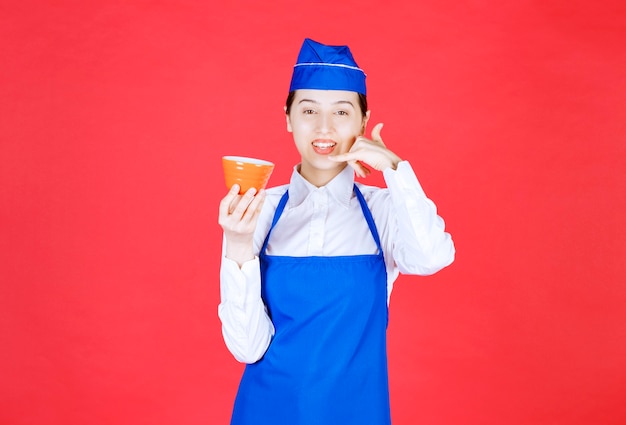 Garçonete de mulher de uniforme segurando uma tigela de laranja e fazendo gesto de telefonema.