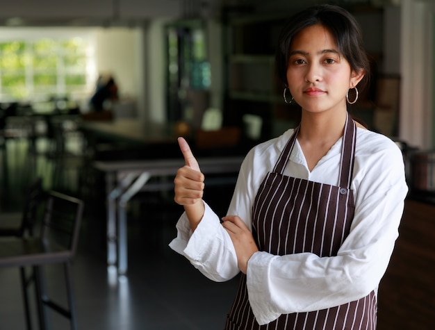 Garçonete feminina étnica confiante no avental em pé com o polegar para cima no café e olhando para a câmera.