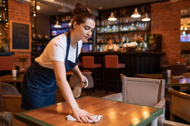 Garçonete muito jovem de avental curvada sobre a mesa de madeira enquanto usa detergente e espanador para limpá-la para os novos clientes do restaurante