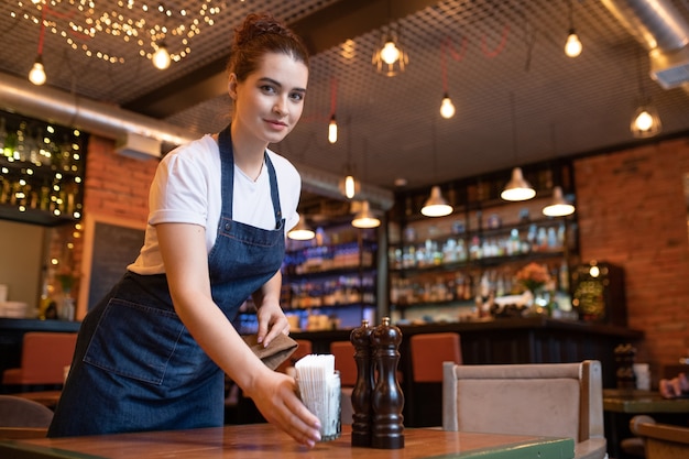 Garçonete muito jovem em trajes de trabalho, curvando-se sobre a mesa enquanto coloca um copo com palitos e olha para você em um café ou restaurante